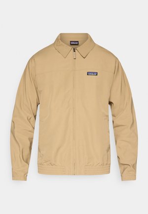 Brun jakke med glidelås foran, spiss krage, elastiske mansjetter og midjebånd. Har en liten blå Patagonia-logo på venstre bryst.