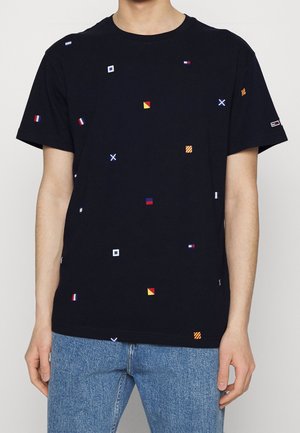 Person iført en sort t-shirt med små farverige nautiske flagmønstre og lyseblå denimjeans, stående foran en ensfarvet baggrund.