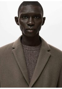 Abrigo de lana marrón con solapa con muesca, usado sobre un suéter de punto texturizado de color marrón oscuro. Tela lisa, diseño minimalista, paleta de colores neutros.