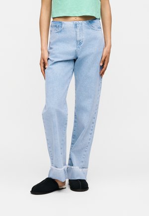 Personne portant un jean bleu clair avec revers, un top court vert menthe et des chaussures slip-on noires avec une bordure blanche, debout devant un fond uni.