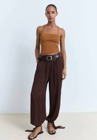 Smeđi pleteni crop top, visoko strukirane smeđe hlače s remenom i vezicama, uparen s crnim sandalama s trakama. Glatka tekstura, opušteni kroj.
