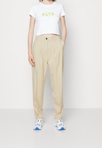 Witte cropped t-shirt met groene spiraalprint, beige plooibroek met zijzakken en witte sneakers met blauwe accenten.