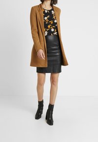 Cappotto di lana marrone sopra un top floreale nero, abbinato a una gonna a matita in pelle nera e stivaletti neri chunky con tacco.