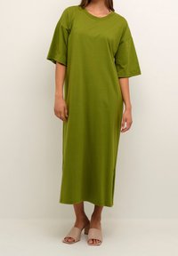 Olivgrön, löst sittande klänning med korta ärmar och slitsar i sidorna, tillverkad av mjuk bomull. Modellen bär beige snörningsskor.