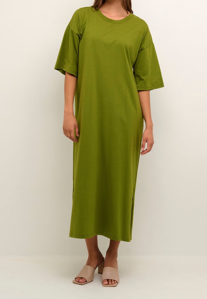 Olivgrön, löst sittande klänning med korta ärmar och slitsar i sidorna, tillverkad av mjuk bomull. Modellen bär beige snörningsskor.