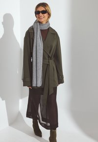 Manteau long vert olive avec une taille ceinturée, associé à une écharpe grise, une robe noire transparente et des bottines vertes. Des lunettes de soleil complètent le look.