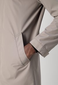 Beige vedenkestävä takki, jossa on taskudetaili, sileä pinta ja muotoiltu istuvuus. Hihansuut ovat hieman istuvat mustalla sisävuorella.