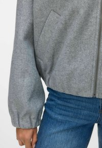 Gros plan sur une personne portant une veste zippée gris clair avec des poignets élastiques et un jean en denim bleu, main détendue le long du corps.