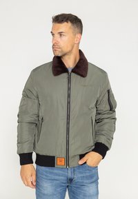 Veste bomber vert olive avec un col en fausse fourrure marron, fermeture éclair, poignets et ourlet côtelés, et une étiquette de marque orange à l'avant.