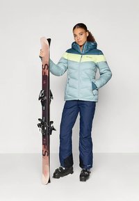 Svijetloplava puffer jakna s tamnoplavo-žutom prugom, uparena s tamnim hlačama, crnim skijaškim čizmama i skijama s ružičastim vrhovima i vezovima.