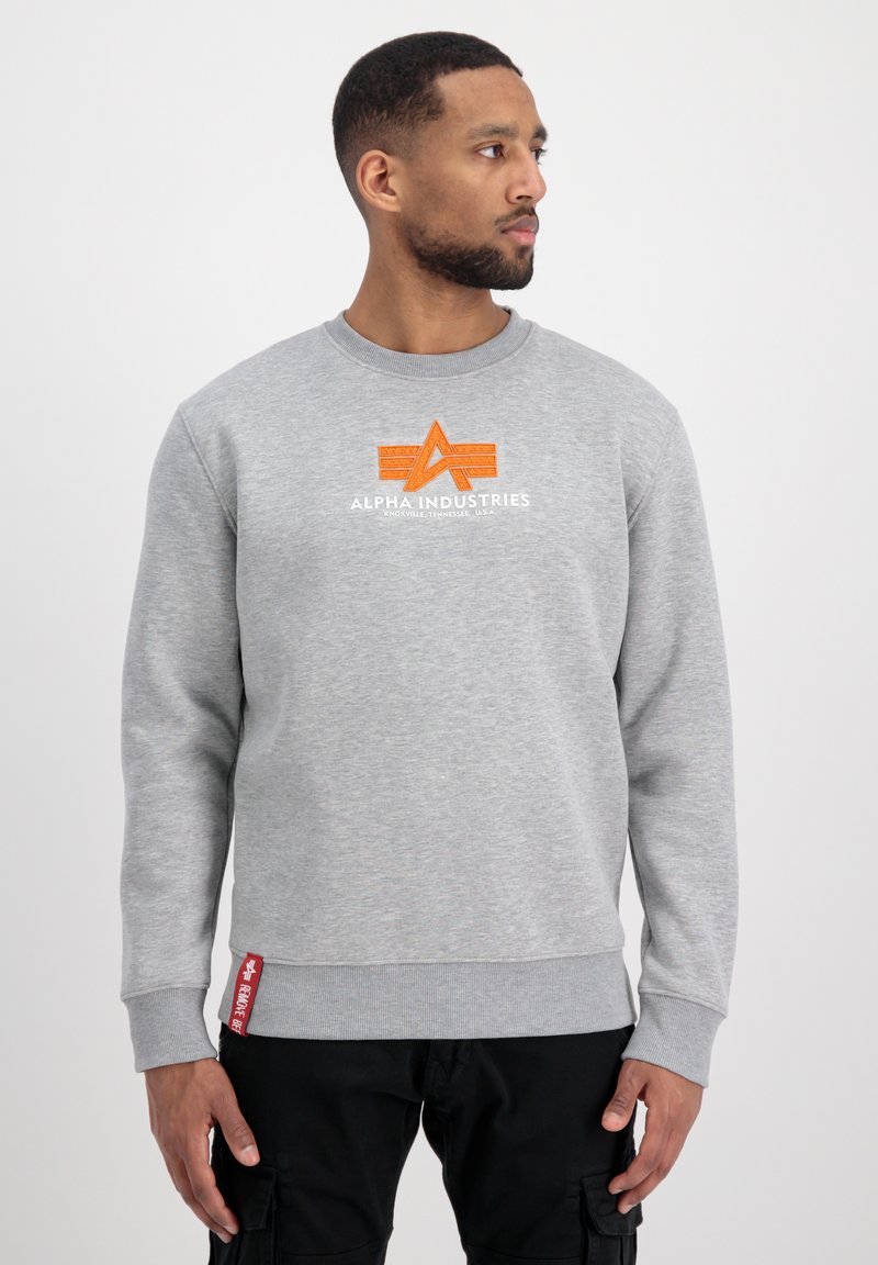 Mężczyzna w szarym swetrze Alpha Industries z pomarańczowym logo, patrzący w prawo, z rękami luźno opadającymi wzdłuż ciała na białym tle.