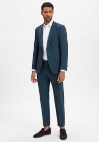 Donkerblauwe suit met een op maat gemaakte pasvorm, gestructureerde stof, met schoudernaden en een witte overhemden. Gecombineerd met marineblauwe loafers.