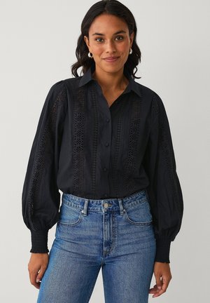 Kvinde iført en sort bluse med lange ærmer med blondeudsnit og blå højtaljede jeans, smilende mod en ensfarvet baggrund.