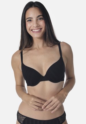 Lächelnde Frau mit langen Haaren, die einen schwarzen BH und passende Spitzenunterwäsche vor einem einfarbigen hellen Hintergrund trägt.