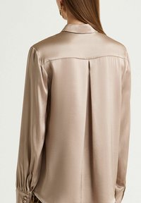 Blusa di seta, colore beige, maniche lunghe con polsini abbottonati, dettaglio piega posteriore, texture liscia, design con colletto classico.