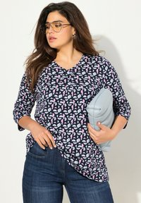 Navybluse mit geometrischen rosa- und weißen Mustern. Hat einen runden Ausschnitt und Dreiviertelärmel. Kombiniert mit blauen Jeans und einer hellgrauen Clutch.