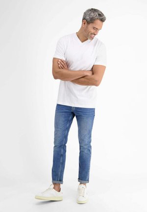 Man with gray hair wearing white t-shirt, blue jeans, and white sneakers stands with arms crossed, smiling and looking down.