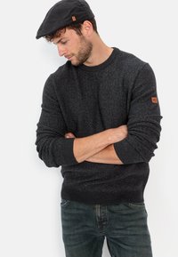 Dunkelgrauer, strukturiertes Strickpullover mit Rundhalsausschnitt und gerippten Bündchen, kombiniert mit dunklen Jeans. Auf dem Ärmel befindet sich ein braunes Logopatch.