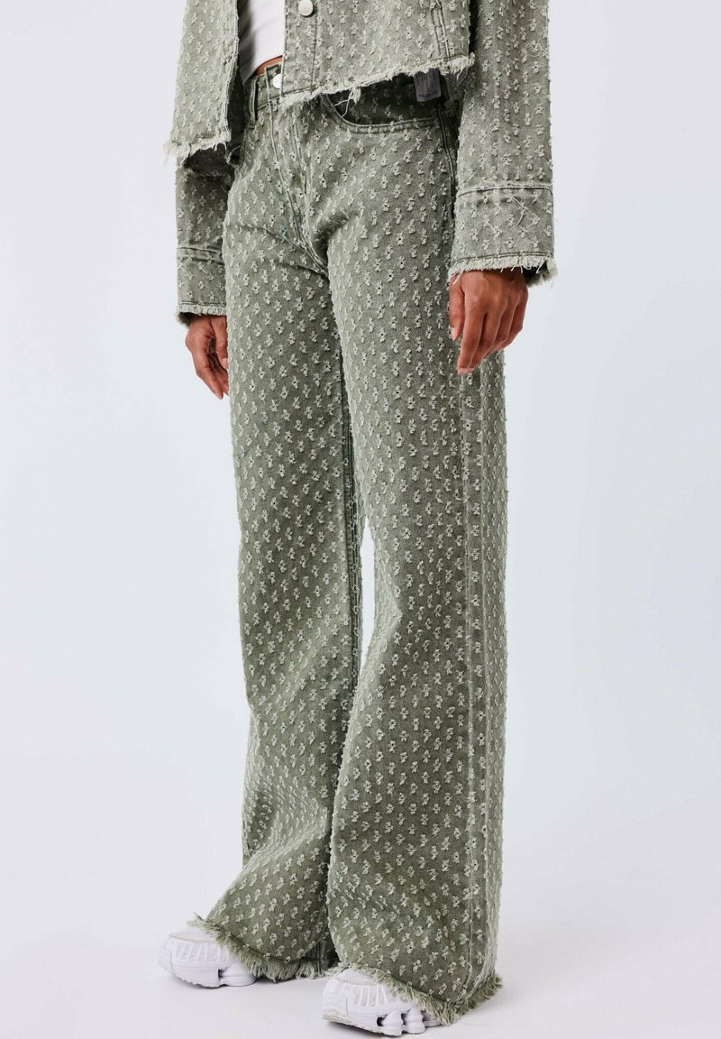 Pantalones de mezclilla acampanados de patrón verde con un acabado texturizado, dobladillos sin terminar y bolsillos laterales, combinados con zapatillas blancas.