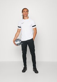 Hvid atletisk T-shirt med sorte striber på ærmerne, parret med sorte slim-fit svedbukser. En sølvfarvet fodbold holdt i den ene hånd.