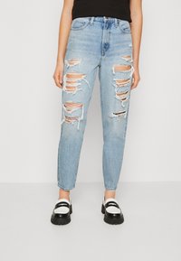 Ljusblå högmidjade rivna jeans med stora slitna fläckar på båda benen, kombinerat med svarta och vita chunky loafers och en svart topp.