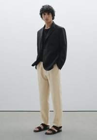 Blazer noir avec deux poches avant, associé à une chemise noire, un pantalon beige et des sandales marron. Tissus lisses, coupe ajustée.