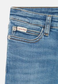 Modré džínsy z denimu s etiketou Calvin Klein, s kontrastným prešívaním a kovovými prvkami na vreckách. Hladká, mierne vyblednutá textúra.