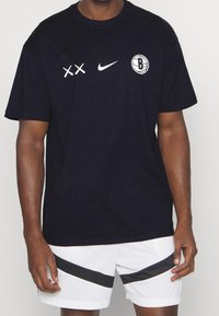 Camiseta de marina de corta manga, con gráficos en blanco: dos X, logo de Nike y un logo circular de Brooklyn en la parte superior izquierda del pecho.
