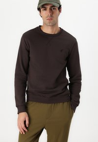Brauner Sweatshirt mit Rundhalsausschnitt, gerippten Bündchen und kleinem gesticktem Logo auf der Brust, kombiniert mit olivgrünen Hosen.