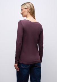 Długą bluzkę w głębokim mauve, wykonaną z miękkiego materiału, z okrągłym dekoltem i dopasowanym krojem. W połączeniu z ciemnoniebieskimi dżinsami, prezentującymi dopasowany widok z tyłu.