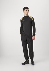 Fekete sportos tréningruha fél cipzáros felsőrésszel és arany kiegészítőkkel, karcsú szabású és rugalmas mandzsettákkal. A megjelenést fekete Nike sportcipő zárja le.
