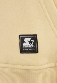 Un tessuto giallo con una trama a coste presenta un'etichetta nera rettangolare su cui è scritto "STARTER BLACK LABEL" in lettere bianche.