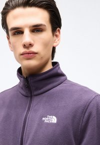 Νέος άνδρας με σκουρόχρωμα μαλλιά φοράει μοβ φλις μπουφάν με φερμουάρ και το λογότυπο The North Face στο στήθος, μπροστά σε μια απλή λευκή φόντο.