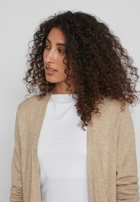 Cardigan beige en tissu doux, à ouverture devant avec des manches longues, porté sur un top blanc, présentant une texture confortable et une coupe décontractée.