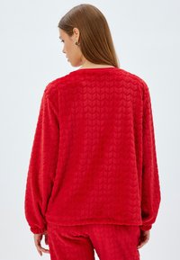 Punainen fleece-paita, jossa on teksturoitu herringbone-kuvio, pitkät hihat ja pyöreä kaula-aukko. Pehmeä, ylellinen kangas ja joustavat hihansuut.