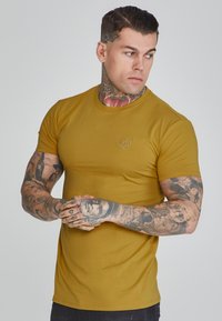Rövid ujjú mustársárga póló, testhez simuló fazonnal, kis hímzett logóval a mellrészen és sima, struktúrált anyaggal.