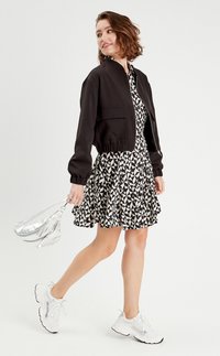 Giacca bomber nera cropped con polsini elastici, indossata sopra un vestito con fantasia in bianco e nero. Accostata a scarpe da ginnastica bianche e una borsa argentata.