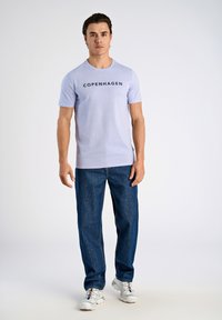 Vaaleanvioletti lyhythihainen t-paita, jossa on teksti "COPENHAGEN", yhdistettynä tummansinisiin suoriin farkkuihin ja valkoisiin tennareihin. Yksinkertainen muotoilu, rento istuvuus.