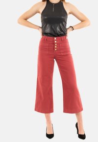 Pantaloni rossi a gamba ampia con cinque bottoni frontali, due tasche anteriori e una superficie liscia, abbinati a un top halter nero lucido e tacchi neri.