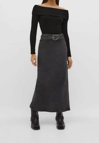 Zwarte off-shoulder geribbelde top gecombineerd met een lange, donkere denimrok; voorzien van een riem met een zilveren gesp en een aansluitende pasvorm. Zwarte enkellaarsjes.