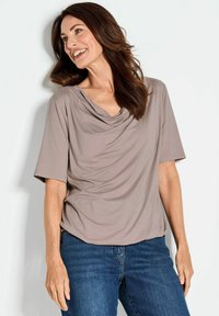 Taupe-farbenes Kurzarmoberteil mit einem schulterfreien Ausschnitt, aus weichem Stoff. Kombiniert mit blauen Jeans, die einen lockeren Schnitt und sanfte Drapierungen aufweisen.