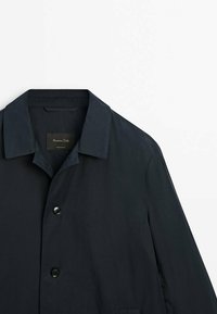 Vaaleansininen kevyt takki, jossa on klassinen kaulus, kaksi nappia ja hienovarainen tekstuuri. Tunnus näkyvissä kaulusalueella.