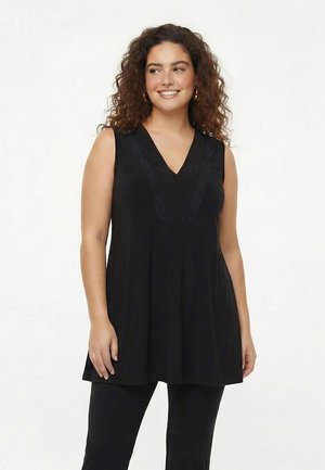 Lächelnde Frau mit lockigem Haar trägt eine ärmellose schwarze Tunika mit V-Ausschnitt und Spitzenbesatz, kombiniert mit schwarzen Hosen, vor einem schlichten Hintergrund.