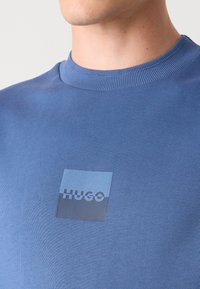 Modrá bavlnená mikina so zaobleným výstrihom a štvorcovou nášivkou s nápisom "HUGO" v čiernej farbe. Hrebienková textúra na golieri.