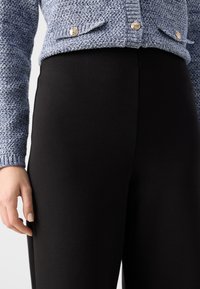 Tkaný modrý cardigan s texturovanou látkou a stříbrnými knoflíky, v kombinaci s obepnutými černými kalhotami. Důraz na ruku spočívající na boku kalhot.