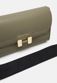 Olivgrön läderclutch med guldfärgade detaljer, som har ett lås. Svart tygrem med präglad text "MAISON." Enkel rektangulär design.