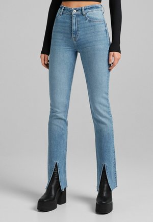 Vrouw draagt lichtblauwe jeans met hoge taille en voorste pijpsplitten, gecombineerd met zwarte plateau-enkellaarzen en een zwart cropped top met lange mouwen.