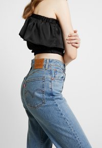 Svart satin off-shoulder crop-top med volangdetaljer, kombinerad med högmidjade ljusblå jeans i denim med en läderlogotyp.
