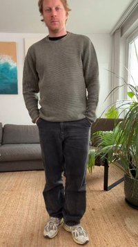 Gray ribbed sweater with long sleeves, paired with dark gray jeans and light sneakers. Interior setting with plants and a couch visible.