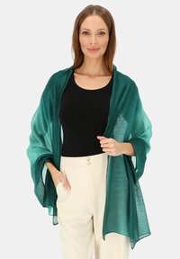 Zielony szal o gradientowym kolorze wykonany z lekkiego, przejrzystego materiału, elegancko układający się na czarnej bluzce i kremowych spodniach. Gładka struktura i miękkie krawędzie.